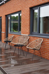 Close up of wicker chairs and sofa with red brick building on the back with glass windows. After the rain with water spots on wooden deck. Summer day. Tallinn, Estonia, Europe. July 2024