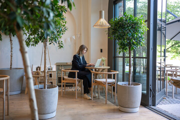 Happy smiling woman employee with laptop in cafe, public space, performing work duties remotely from comfortable quiet cozy place. Satisfied female attentively, seriously doing job at restaurant table