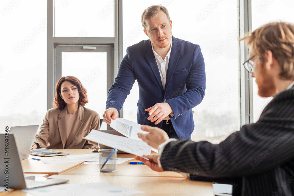 Poster a businessman in a blue suit stands and hands documents to a seated colleague while another sits in 