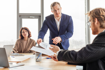 A businessman in a blue suit stands and hands documents to a seated colleague while another sits in the background, all gathered around a conference table in an office setting.