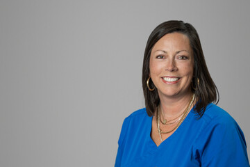 Smiling headshot of Caucasian brunette middle age medical worker woman in a blue scrubs top in a studio on gray grey backdrop