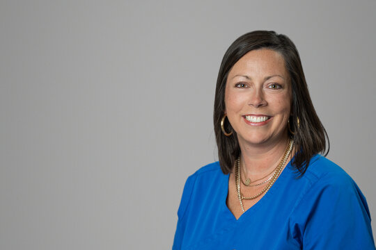 Smiling headshot of Caucasian brunette middle age medical worker woman in a blue scrubs top in a studio on gray grey backdrop