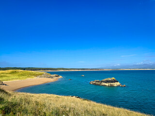 Obraz premium A serene beach in Anglesey, encircled by majestic cliffs, where golden sands meet turquoise waters under a vast, clear sky. Perfect natural beauty.
