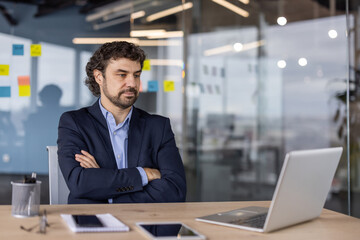 Fototapeta premium Business professional in modern office working on laptop, tablet, and phone on desk. Serious expression with arms crossed, surrounded by sticky notes and glass walls.