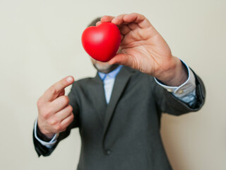 Red heart in businessman hand in focus, the man is out of focus, light background. Expression of love towards customers of life concept. Model in grey suit and blue office shirt. Positive motivation