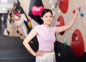 Young Asian girl poses near steep climbing wall in gym and leans against hook. Woman is resting...