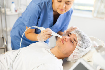 Young woman cosmetologist performs facial exfoliation procedure with apparatus young male patient