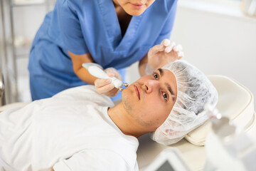 Young female cosmetologist performs hardware facial microcurrent procedure to young male patient