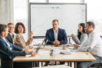 A group of business professionals are gathered around a table in a modern office, discussing a growth chart displayed on a large screen. The team is engaged in a lively conversation