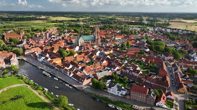 Aerial shot of historic town Ribe with majestic Ribe cathedral, red-roofed buildings and boat moving along the river, set against lush, green landscape, south-west Jutland, Denmark.