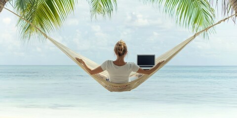 African American woman working on a laptop in a hammock by the tropical beach, palm trees and turquoise sea in the background. Remote work concept.
