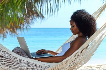African American woman working on a laptop in a hammock by the tropical beach, palm trees and turquoise sea in the background. Remote work concept.
