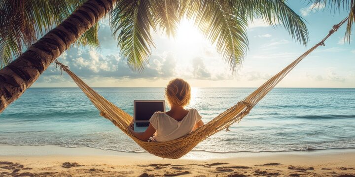 African American woman working on a laptop in a hammock by the tropical beach, palm trees and turquoise sea in the background. Remote work concept. - Powered by Adobe