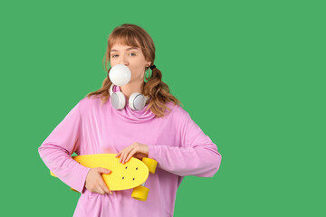 Beautiful young woman with chewing gum and skateboard on green background