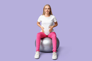 Beautiful young sporty pregnant woman with fitball on lilac background