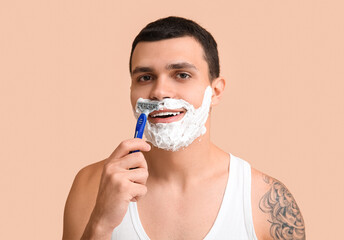 Handsome young man with razor shaving his face on beige background