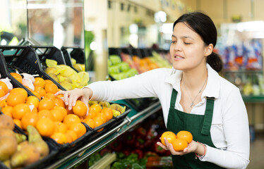 Naklejka premium Young woman seller in uniform sells tangerines in grocery store..