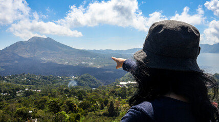 Back view of a female tourist standing on a viewpoint pointing at Mount Batur. Volcanic landscape and surrounding greenery are visible under a clear sky, ideal for travel and adventure related content