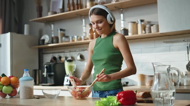 Earphones lady making salad in cozy kitchen dancing to music. Woman preparing