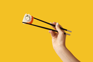 Female hand holding chopsticks with tasty sushi roll on yellow background