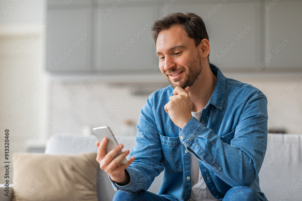 Poster a man with a beard sits on a couch and looks at a smartphone with a smile on his face. he is wearing