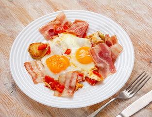 Delicious homemade breakfast of fried eggs with potatoes, pieces of ripe tomatoes and crispy streaky side bacon slices..