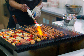 Close up of a chef's hands grilling sausage skewers on a BBQ, with shrimp and vegetables sizzling on the hot grill. with fire