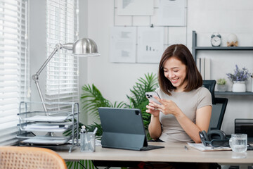 A young woman in a home office setting, smiling while using her smartphone. The workspace includes a laptop, documents, and modern decor.