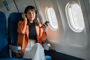 A young woman wearing headphones and holding a smartphone, sitting comfortably in an airplane seat by the window.