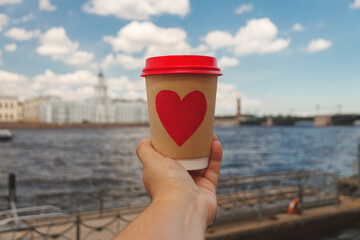 Brown paper cup of natural coffee with red heart in woman's palm near embankment of Neva river in St. Petersburg