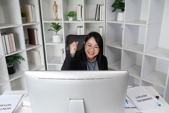Successful Businesswoman Working on Computer in Modern Office with Bookshelves and Plants, Celebrating Achievement