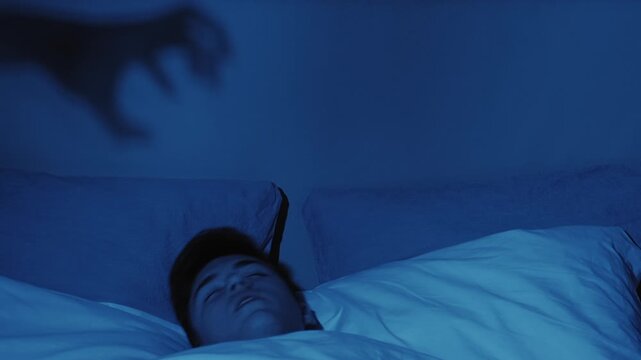 Bad dream. Night horror. Sleep anxiety. Frightened man woke up in bed from nightmare of creepy evil demon hand shadow in dark bedroom.