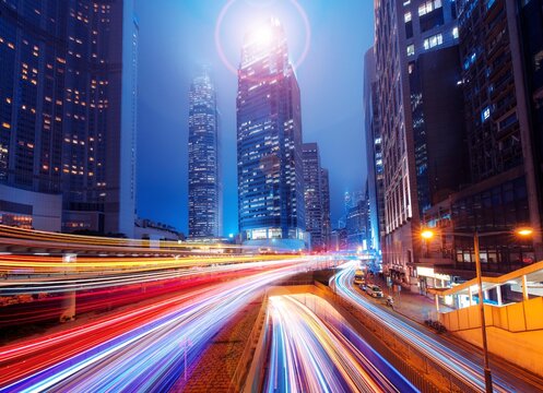 A night scene of a Hong Kong street with tall buildings and brightly lit windows. Light trails from cars moving along the street create a colorful and dynamic scene.
