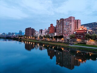 A tranquil waterfront cityscape at dusk with illuminated buildings reflecting in the calm river....