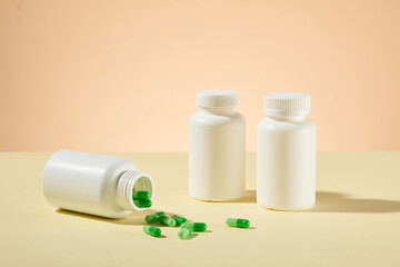 Light yellow countertop displaying some mockup medicine bottles for adding brand's name, one flat lay and pouring green capsule onto table, others standing. Front view, empty space for designing