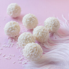 Top view of White Chocolate Candy With Coconut Topping On White Feather and pink background, sweet balls on plate flat lay with copy space. Romantic gift for celebration or anniversary