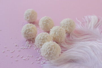 Top view of White Chocolate Candy With Coconut Topping On White Feather and pink background, sweet balls on plate flat lay with copy space. Romantic gift for celebration or anniversary