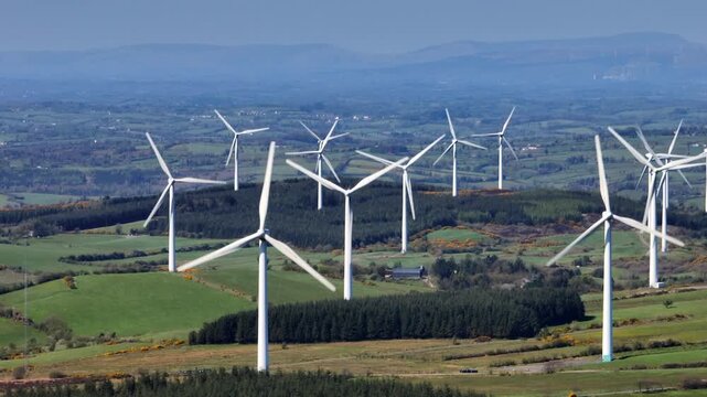 Wind Turbines, Cornasaus, County Cavan, Ireland, April 2023. Drone tracks slowly with a Telephoto lens as windmills rotate above green farmland with the mountains of Ulster in the distance.