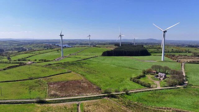 Wind Turbines, Cornasaus, County Cavan, Ireland, April 2023. Drone orbits counter clockwise as windmill blades rotate over beautiful green farmland under a clear blue sky.