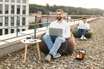 Business man sitting on bean bag chair and connects charging cable to portable power station on...