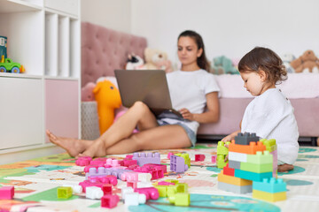 cute little baby girl playing with toys blocks and working mother sitting on floor with laptop in room