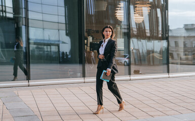 Elegant businesswoman in a black suit walking briskly outside a glass office building, holding a...