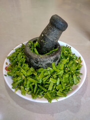 A stone mortar and pestle filled with finely chopped green cluster beans rests on a white plate.