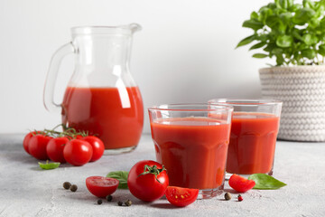 Tasty tomato juice in glasses, jug, basil, peppercorns and fresh vegetables on light grey table
