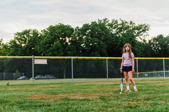 A serious girl stands in field with tennis racket