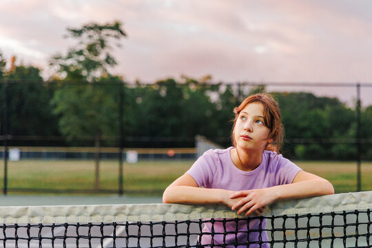 A girl looks leans on net at tennis court at sunset looking up