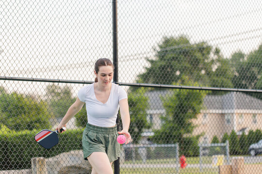 Young woman playing pickleball with a pink ball on an outdoor court.
