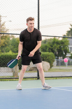 Young man playing pickleball with a pink ball on an outdoor court.