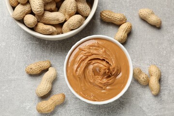 Tasty peanut butter in bowl and groundnuts on grey textured table, flat lay