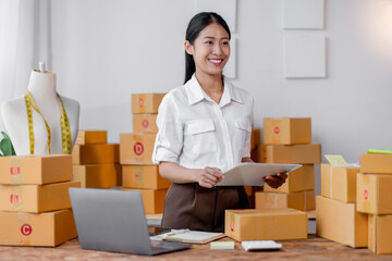 Startup SME small business asian entrepreneur SME or freelance Asian woman standing using a laptop,clipboard with box, online marketing packaging box and delivery, SME concept.